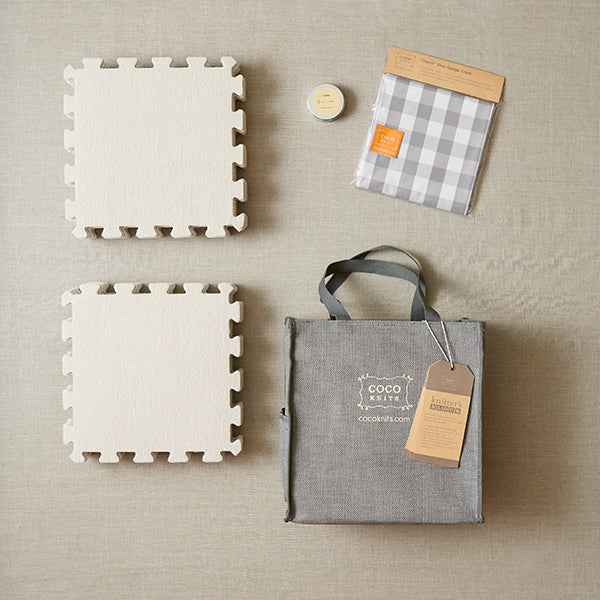 A top view of the CocoKnits Knitter,s Block kit with the items displayed: a tin of 40 1.5 inch stainless steel T-pins, 18 12x12 inch interlocking fiber-topped foam blocking mat squares, 48 inch cotton cloth with 1 inch gingham squares, and a jute carrying bag 